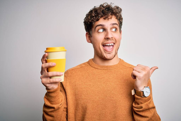 Nuori vaalea mies, jolla on kiharat hiukset juominen kuppi kahvia seisoo valkoisen taustan yläpuolella osoittaen ja osoittaen peukalolla sivulle hymyillen
 - Valokuva, kuva