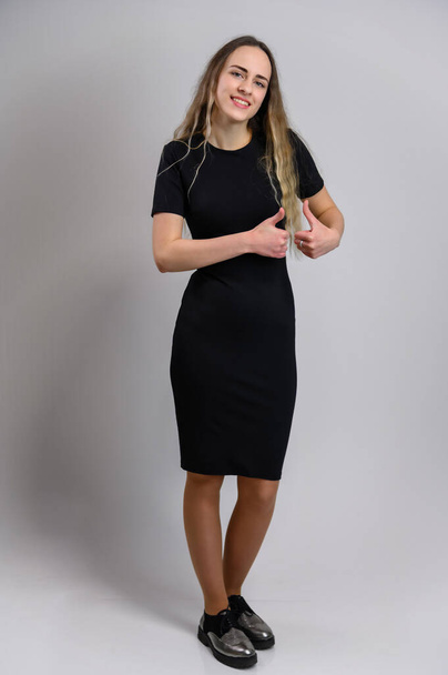 Vertical photo in full on a white photo growth Caucasian model with long hair in a black dress. The girl is standing upright, showing ok with her hands with a smile. - Photo, Image