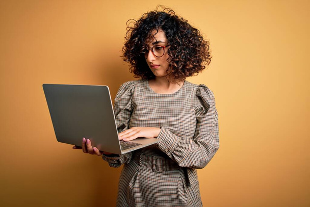 Beautiful arab businesswoman wearing glasses working using laptop over yellow background with a confident expression on smart face thinking serious - Valokuva, kuva
