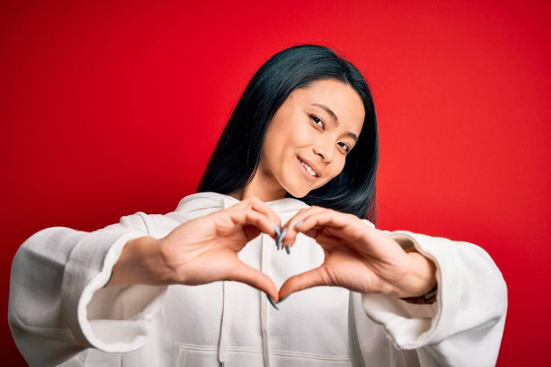 Jovem mulher esportiva chinesa bonita vestindo camisola sobre fundo vermelho isolado sorrindo no amor fazendo forma de símbolo do coração com as mãos. Conceito romântico
. - Foto, Imagem