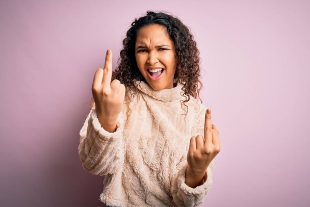 Young beautiful woman with curly hair wearing casual sweater standing over pink background Showing middle finger doing fuck you bad expression, provocation and rude attitude. Screaming excited - Fotografie, Obrázek