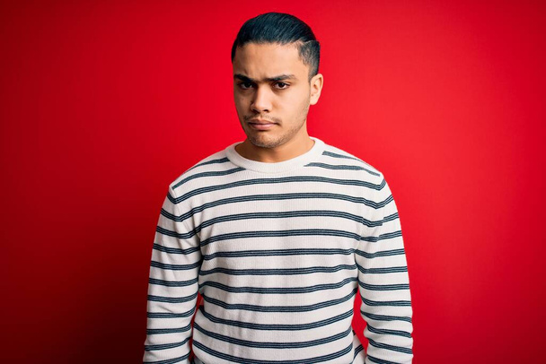 Mladý brazilský muž v neformálním pruhovaném tričku, stojící nad izolovaným červeným pozadím, skeptický a nervózní, a mračí se, protože má problém. Negativní osoba. - Fotografie, Obrázek