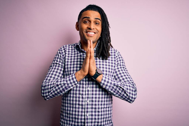 Молодой красивый африканский афроамериканец с дредами в повседневной рубашке молится руками, прося прощения, уверенно улыбаясь.
. - Фото, изображение