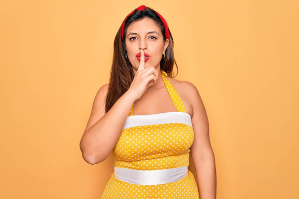 Junge hispanische Pin-up-Frau trägt Mode sexy 50er Jahre Stil vor gelbem Hintergrund und bittet um Ruhe mit dem Finger auf den Lippen. Schweigen und geheimes Konzept. - Foto, Bild
