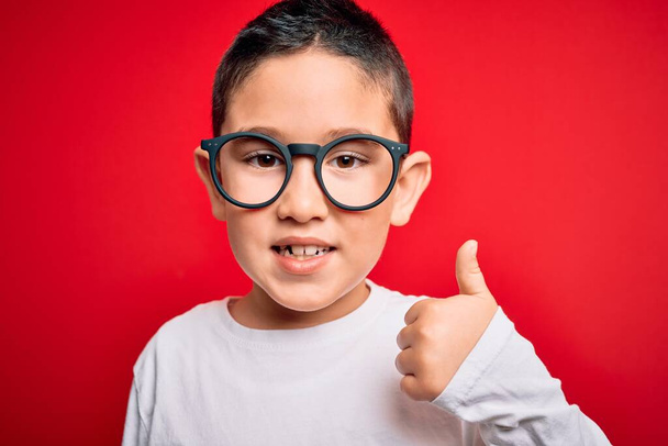 Mladý malý chytrý chlapec nosí nerd brýle přes červené izolované pozadí šťastný s velkým úsměvem dělá ok znamení, palec nahoru s prsty, vynikající znamení - Fotografie, Obrázek