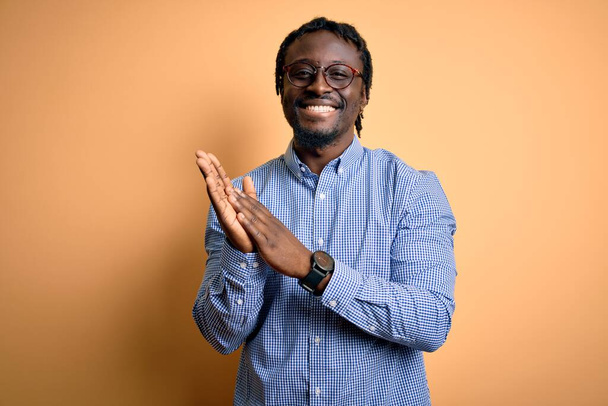 Junger gutaussehender afrikanisch-amerikanischer Mann mit Hemd und Brille vor gelbem Hintergrund klatscht und applaudiert fröhlich und fröhlich, lächelt stolz in die Hände - Foto, Bild