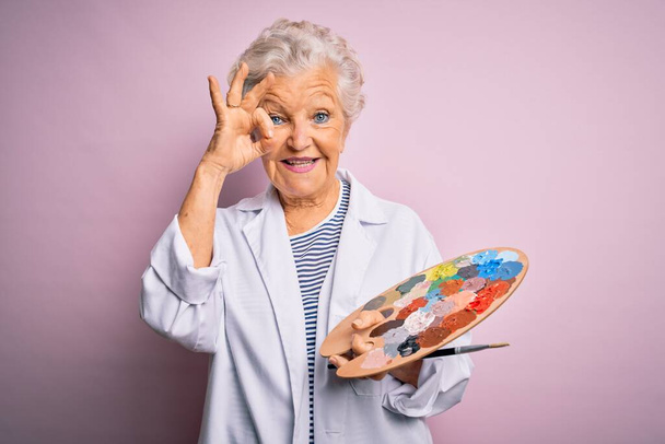 Старша красива жінка-художниця, яка малює пензлем і палітрою на рожевому тлі, робить знак з пальцями, відмінний символ
 - Фото, зображення