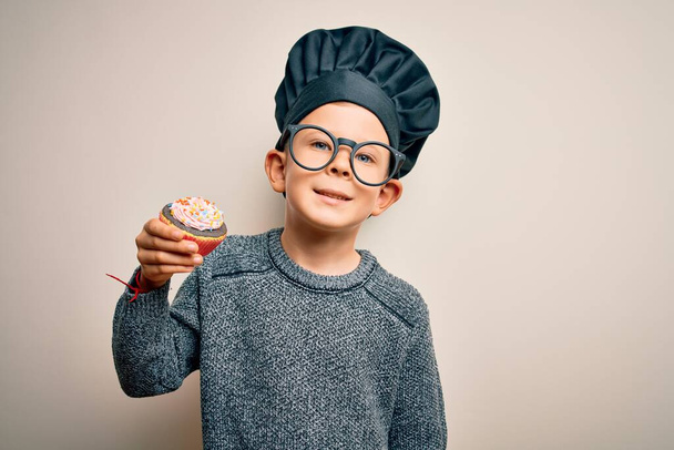 シェフの制服を着た若い白人料理人の子供と幸せな顔をした帽子調理カップケーキ歯を示す自信を持って笑顔で立って笑顔 - 写真・画像