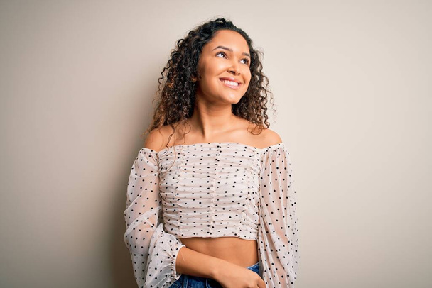 Young beautiful woman with curly hair wearing casual t-shirt standing over white background looking away to side with smile on face, natural expression. Laughing confident. - Foto, afbeelding