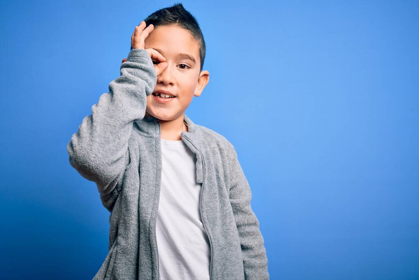 Νεαρό μικρό παιδί φορώντας αθλητική μπλούζα πάνω από το μπλε απομονωμένο φόντο κάνει εντάξει χειρονομία με το χέρι χαμογελώντας, το μάτι κοιτάζοντας μέσα από τα δάχτυλα με χαρούμενο πρόσωπο. - Φωτογραφία, εικόνα