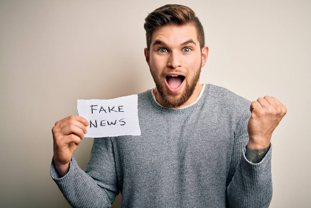 Young blond man with beard and blue eyes holding paper with fake news message screaming proud and celebrating victory and success very excited, cheering emotion - Photo, Image