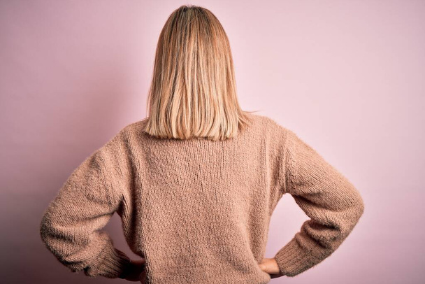 Junge schöne blonde Frau trägt winterlichen Wollpullover über rosa isoliertem Hintergrund, steht rückwärts und blickt mit den Armen am Körper weg - Foto, Bild