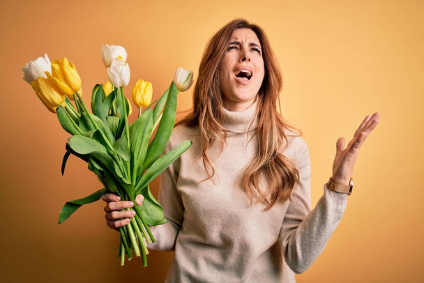 Young beautiful brunette woman holding bouquet of yellow tulips over isolated background crazy and mad shouting and yelling with aggressive expression and arms raised. Frustration concept. - Photo, Image