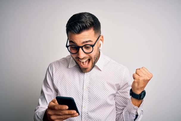 Young handsome man wearing glasses having conversation using smartphone and earphones screaming proud and celebrating victory and success very excited, cheering emotion - Photo, image