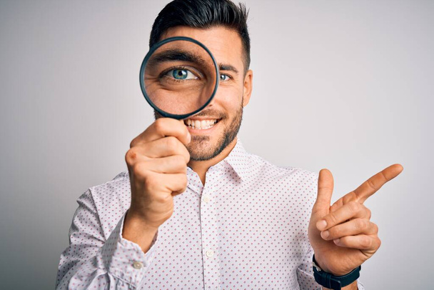 Young detective man looking through magnifying glass over isolated background very happy pointing with hand and finger to the side - Foto, Bild