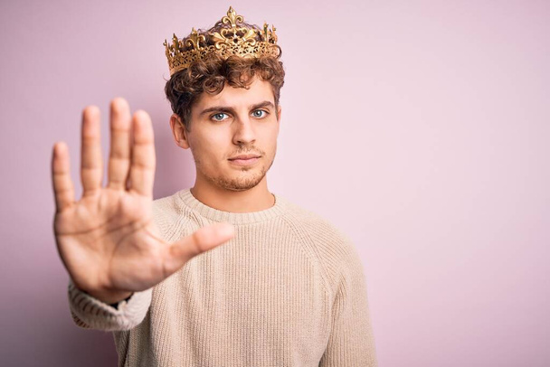 Young blond man with curly hair wearing golden crown of king over pink background doing stop sing with palm of the hand. Warning expression with negative and serious gesture on the face. - Foto, Bild