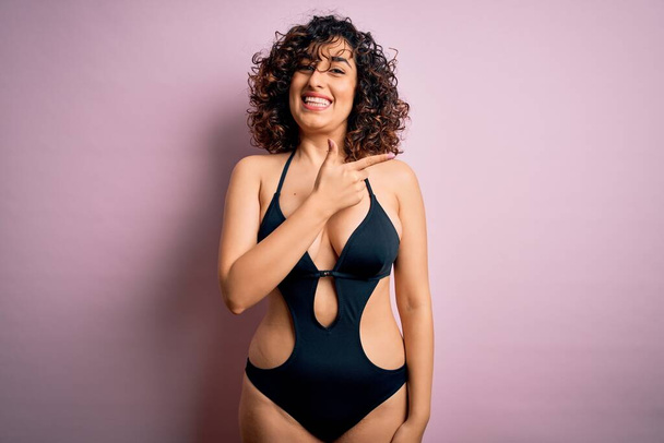 Young beautiful arab woman on vacation wearing swimsuit and sunglasses over pink background cheerful with a smile of face pointing with hand and finger up to the side with happy and natural expression on face - Фото, зображення