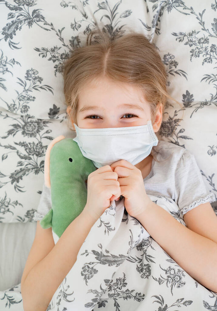 girl lying in bed in medical mask. dinosaur - Zdjęcie, obraz