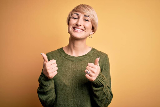 Jonge blonde vrouw met modern kort haar draagt casual trui over gele achtergrond succes teken doet positieve gebaar met de hand, duimen omhoog glimlachend en gelukkig. Vrolijke uitdrukking en winnaar gebaar. - Foto, afbeelding