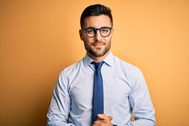 Young handsome businessman wearing tie and glasses standing over yellow background with hands together and crossed fingers smiling relaxed and cheerful. Success and optimistic - Foto, Imagen