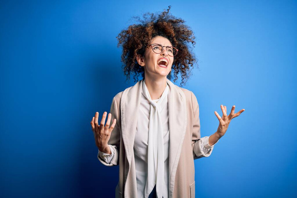 Young beautiful businesswoman with curly hair and piercing wearing jacket and glasses crazy and mad shouting and yelling with aggressive expression and arms raised. Frustration concept. - Φωτογραφία, εικόνα