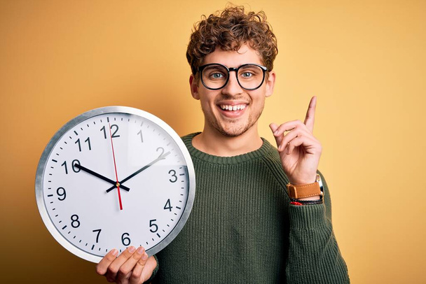 Junger blonder Mann mit lockigem Haar, Brille, große Uhr vor gelbem Hintergrund, überrascht mit einer Idee oder Frage, Zeigefinger mit glücklichem Gesicht, Nummer eins - Foto, Bild