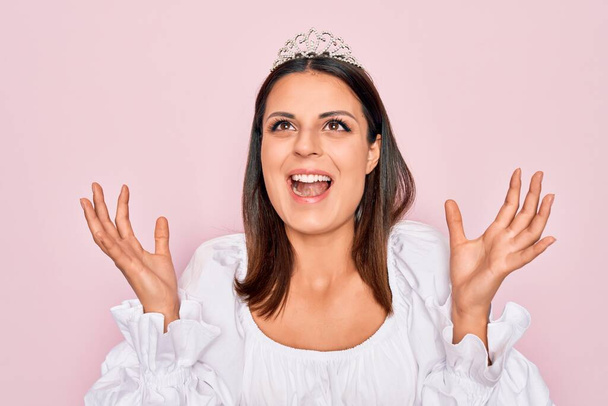 Young beautiful brunette woman wearing princess tiara standing over isolated pink background crazy and mad shouting and yelling with aggressive expression and arms raised. Frustration concept. - Fotoğraf, Görsel