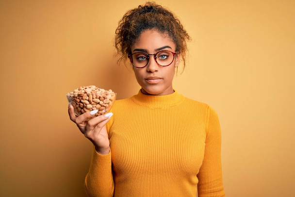 Молодая африканская афроамериканка держит миску со здоровым арахисом на желтом фоне с уверенным выражением на умной морде, думая серьезно
 - Фото, изображение