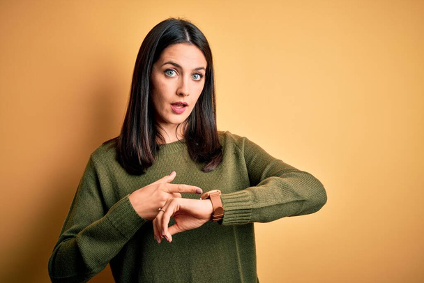 Young brunette woman with blue eyes wearing green casual sweater over yellow background In hurry pointing to watch time, impatience, upset and angry for deadline delay - Fotoğraf, Görsel