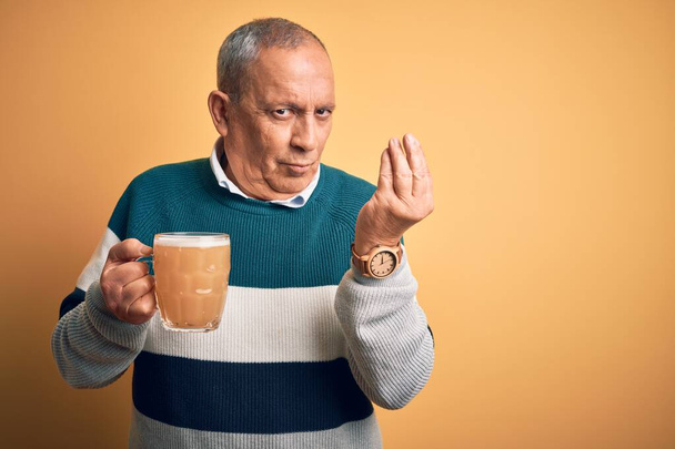 Starszy przystojny mężczyzna pije słoik piwa stojący nad odizolowanym żółtym tłem Wykonując włoski gest z ręką i palcami pewny siebie wyraz twarzy - Zdjęcie, obraz