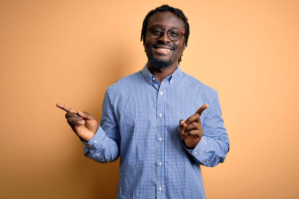 Ein junger gutaussehender afrikanisch-amerikanischer Mann mit Hemd und Brille vor gelbem Hintergrund lächelt selbstbewusst und zeigt mit den Fingern in verschiedene Richtungen. Kopierfläche für Werbung - Foto, Bild