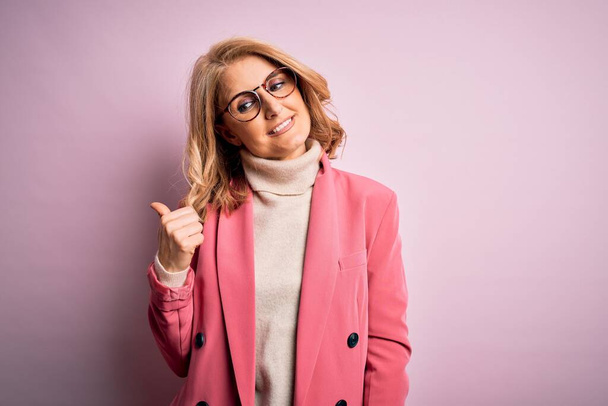 Mulher de negócios loira de meia idade bonita vestindo jaqueta rosa elegante e óculos sorrindo com o rosto feliz olhando e apontando para o lado com o polegar para cima
. - Foto, Imagem