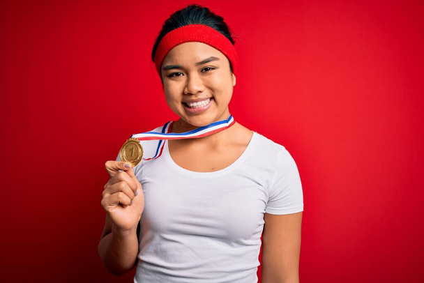 Joven campeona asiática chica ganadora medalla de pie sobre fondo rojo aislado con una cara feliz de pie y sonriendo con una sonrisa confiada mostrando los dientes
 - Foto, Imagen