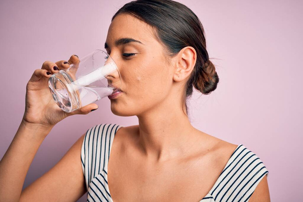 Jovem bela mulher morena bebendo copo de água saudável para refrescar em pé sobre fundo rosa isolado
 - Foto, Imagem
