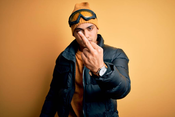Young brazilian skier man wearing snow sportswear and ski goggles over yellow background Showing middle finger, impolite and rude fuck off expression - Foto, Bild