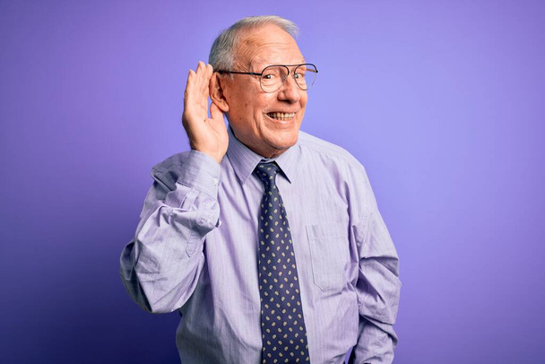Grauhaariger älterer Geschäftsmann mit Brille vor violettem Hintergrund, der lächelnd mit der Hand über dem Ohr Gerüchten oder Klatsch lauscht. Konzept der Gehörlosigkeit. - Foto, Bild