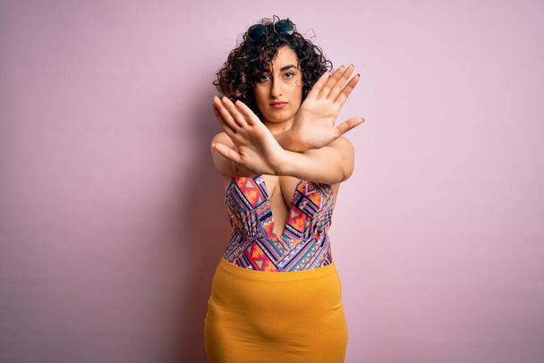 Young beautiful arab woman on vacation wearing swimsuit and sunglasses over pink background Rejection expression crossing arms and palms doing negative sign, angry face - Photo, Image