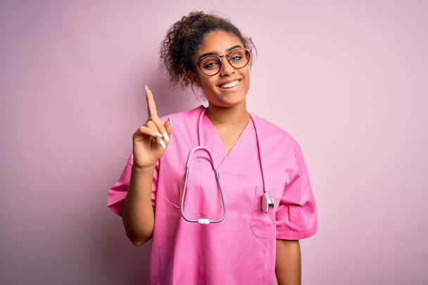 Niña enfermera afroamericana vistiendo uniforme médico y estetoscopio sobre fondo rosa mostrando y señalando hacia arriba con el dedo número uno mientras sonríe confiado y feliz
. - Foto, imagen