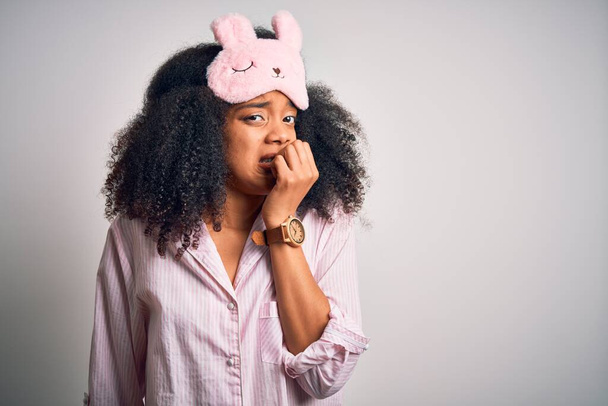 Nuori afroamerikkalainen nainen, jolla on afrokarvat, nukkuva silmänaamio ja vaaleanpunainen pyjama, joka näyttää stressaantuneelta ja hermostuneelta kädet suulla pureen kynsiä. Ahdistuneisuusongelma
. - Valokuva, kuva