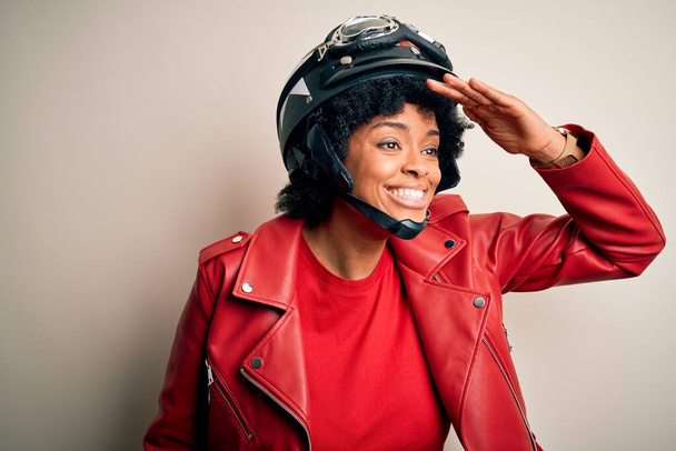 Junge afroamerikanische Motorradfahrerin mit lockigem Haar, Motorradhelm, sehr glücklich und lächelnd, die mit der Hand über dem Kopf in die Ferne blickt. Suchkonzept. - Foto, Bild