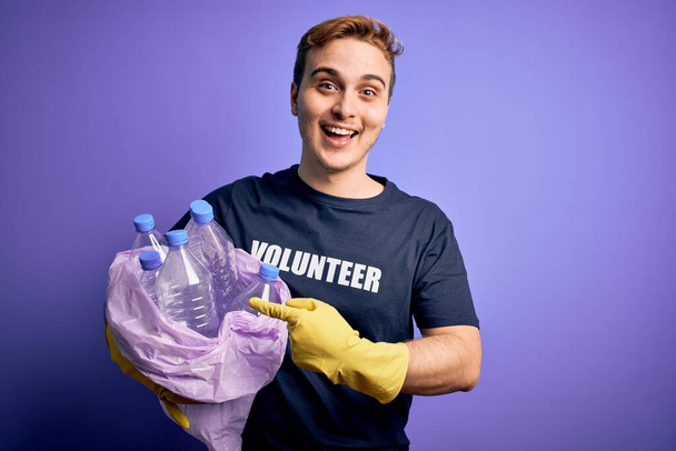 Mladý pohledný zrzka dobrovolník muž dělá dobrovolnické držení taška s plastovými lahvemi Usmívá šťastný ukazující rukou a prstem - Fotografie, Obrázek