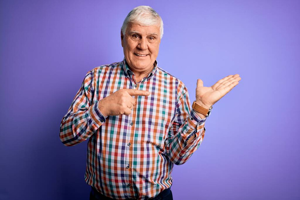 Senior bonito hoary homem vestindo casual camisa colorida sobre isolado roxo fundo espantado e sorrindo para a câmera ao apresentar com a mão e apontando com o dedo
. - Foto, Imagem