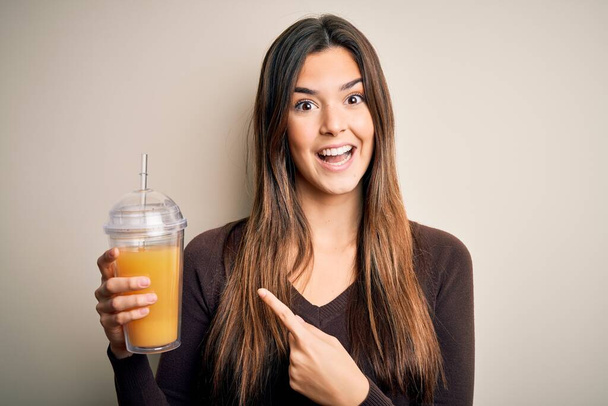 Joven hermosa chica bebiendo vaso de jugo de naranja saludable sobre fondo blanco aislado muy feliz señalando con la mano y el dedo
 - Foto, Imagen