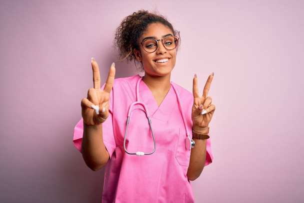 Африканська медсестра з Америки, одягнена в медичну форму і стетоскоп над рожевим фоном, посміхається, дивлячись на камеру з пальцями, які роблять знак перемоги. Номер два. - Фото, зображення