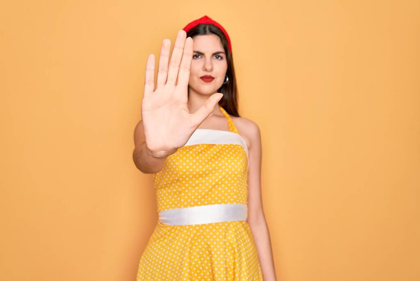 Young beautiful pin up woman wearing 50s fashion vintage dress over yellow background doing stop sing with palm of the hand. Warning expression with negative and serious gesture on the face. - Photo, Image