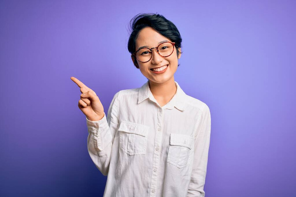 Fiatal gyönyörű ázsiai lány, alkalmi inget és szemüveget visel, lila háttér felett áll, ujjal mutogatva a sikeres ötlettel. Kilépett és boldog. Egyes számú.. - Fotó, kép