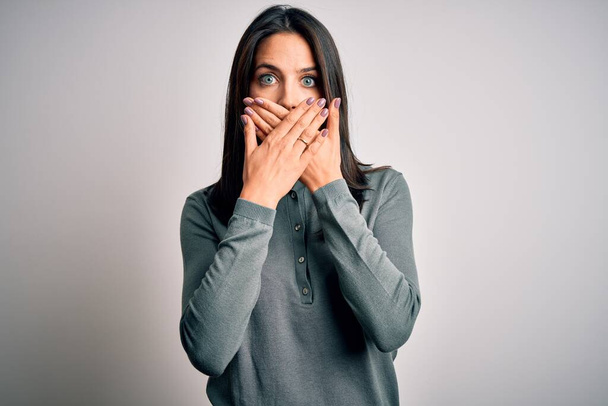 Young brunette woman with blue eyes wearing casual green sweater over white background shocked covering mouth with hands for mistake. Secret concept. - Foto, afbeelding
