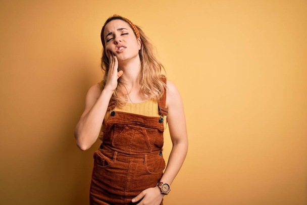 Young beautiful blonde woman wearing overalls and diadem standing over yellow background touching mouth with hand with painful expression because of toothache or dental illness on teeth. Dentist - Foto, immagini