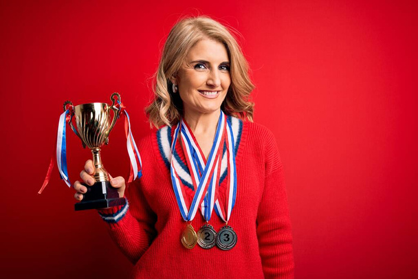 Middle age beautiful blonde successful woman wearing medals holding trophy with a happy face standing and smiling with a confident smile showing teeth - Photo, image