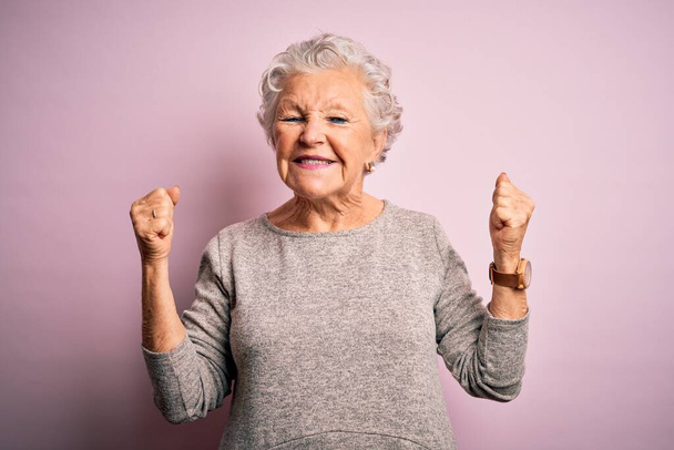 Senior kaunis nainen päällään rento t-paita seisoo yli eristetty vaaleanpunainen tausta innoissaan menestystä kädet esille ja silmät kiinni juhlii voittoa hymyillen. Voittajan käsite
. - Valokuva, kuva